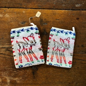 May include: Two rectangular washcloths with a white background, featuring a colorful deer design. The deer is outlined in brown and filled with red and orange. The edges are decorated with blue, green, and red floral patterns.