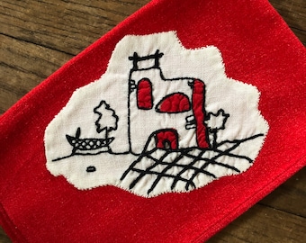 May include: Red fabric kitchen towel with a white embroidered design of a house with a red roof and a black fence.
