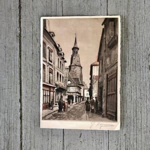 May include: A vintage postcard featuring a sepia-toned illustration of a narrow European cobblestone street lined with multi-story buildings and a prominent central clock tower. Several figures walk along the street. The signature J. Degraven is visible.