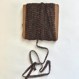 May include: A roll of brown beaded trim with a zigzag pattern. The trim is on a cardboard spool, with a length of the trim unraveled. The trim has a scalloped edge and is a dark, metallic brown color.