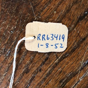 May include: A small, rectangular tag made of light brown cardboard, attached to a white string. The tag has the text "RR63419" and "1-8-52" written in blue ink. The tag is set against a wooden surface.