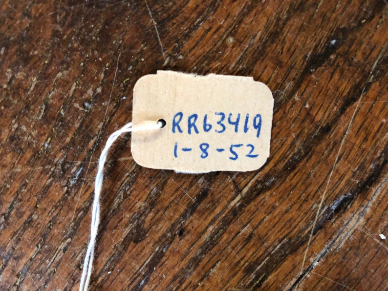May include: A small, rectangular tag made of light brown cardboard, attached to a white string. The tag has the text "RR63419" and "1-8-52" written in blue ink. The tag is set against a wooden surface.