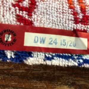 May include: Close-up of a colorful towel with a red tag. The tag has the text "DW 24 15/20" in blue and a logo. The towel has white, red, blue, and orange patterns.
