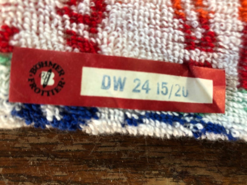 May include: Close-up of a colorful towel with a red tag. The tag has the text "DW 24 15/20" in blue and a logo. The towel has white, red, blue, and orange patterns.