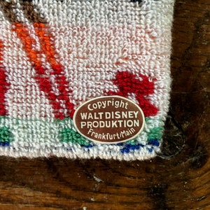 May include: Close-up of a white terry cloth towel with colorful embroidered details. The towel features red, orange, and green accents, with a brown and white label that reads "Copyright Walt Disney Produktion Frankfurt/Main."