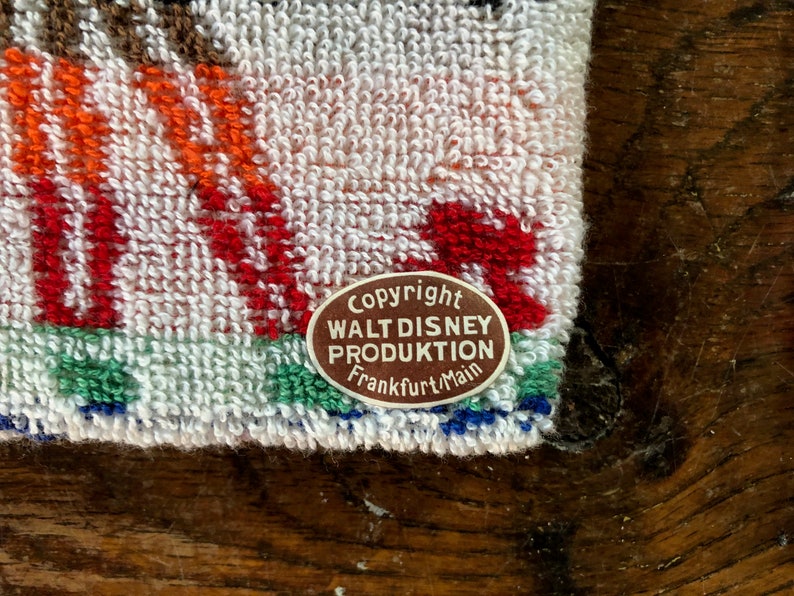 May include: Close-up of a white terry cloth towel with colorful embroidered details. The towel features red, orange, and green accents, with a brown and white label that reads "Copyright Walt Disney Produktion Frankfurt/Main."