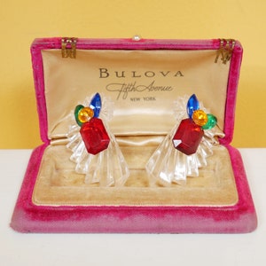 May include: A pair of clear crystal earrings with red, blue, green, and yellow stones in a pink velvet jewelry box. The box has the text "Bulova Fifth Avenue New York" on the inside lid.