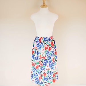 May include: A midi-length skirt with a floral pattern, shown on a mannequin. The skirt has a white base, adorned with red, blue, green, and black flower designs. It features an elasticated waistband.
