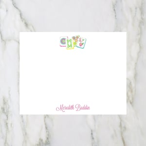 May include: White card with a colorful graphic of the word "HOME" in a block font with flowers and swirls. The name "Meredith Buddin" is printed in pink cursive at the bottom of the card.