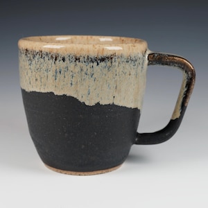 May include: A handmade ceramic mug with a unique design. The mug features a two-tone color scheme, with a black base and a beige top with brown accents. The handle is also brown. The mug is perfect for coffee or tea.