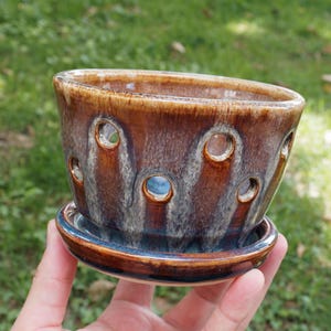 May include: A brown and blue glazed ceramic orchid pot with a matching saucer. The pot features a series of circular cutouts around the sides, allowing for airflow. The glaze has a glossy finish with blended earthy tones of brown, tan, and blue.