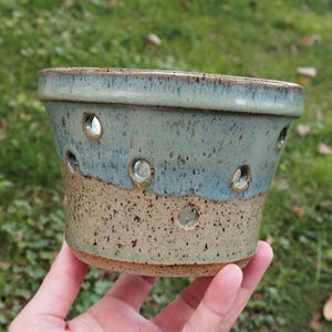 May include: A ceramic planter with a tapered cylindrical shape, featuring a two-tone glaze. The upper portion is a smooth, glossy sage green, while the lower section is a speckled tan. Multiple circular drainage holes are spaced around the body.