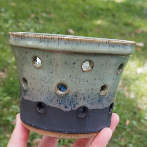 May include: A tapered ceramic planter featuring a two-tone glaze with a speckled sage green top transitioning into a solid matte black base. The vessel is punctuated by multiple circular drainage holes arranged in a staggered pattern around the sides.