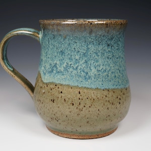 May include: Handmade ceramic mug with a large handle. The mug features a two-tone design, with a textured blue glaze on the upper half and a speckled tan glaze on the lower half. The rim and handle have a darker brown glaze.