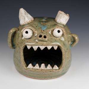 May include: A ceramic monster-faced container with a wide open mouth and two white horns. The container is primarily green with blue and white accents. The monster has large, round eyes and small ears.