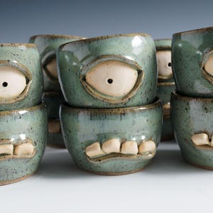 May include: A collection of ceramic mugs featuring a speckled sage green glaze. Each mug is designed with a single large eye and a row of white teeth protruding from the surface, creating a creature-like appearance with a glossy finish.