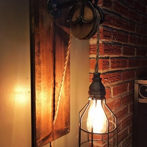 May include: A rustic wall-mounted light fixture with a wooden plank backing. The fixture features a pulley system with a rope and a caged light bulb. The wood is stained a warm brown, and the metal components are black.