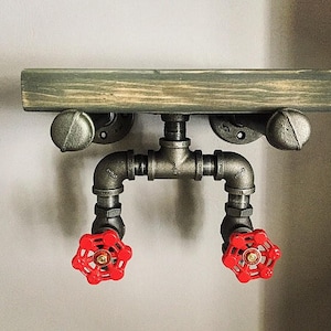 May include: Industrial-style wall shelf featuring a distressed wooden plank supported by galvanized steel pipes and fittings. The shelf includes two red valve handles, adding a pop of color to the design. The shelf is approximately 24 inches wide.