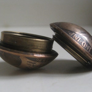 May include: A small, round, metallic container crafted from two halves of a vintage coin. The exterior shows the year 1896 and the text IND IMP. The two pieces are joined by a threaded brass-coloured interior rim, allowing the container to open.