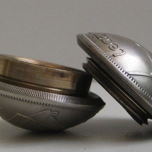 May include: A silver-toned metal container composed of two circular, dome-shaped halves that screw together. The exterior features embossed text and decorative patterns, while the interior reveals a plain, cylindrical brass-coloured metal rim.