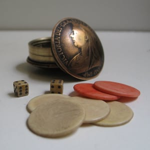 May include: An antique game set featuring a copper coin lid with a profile, a small brass container, two dice, and several game chips in shades of beige and red. The set is likely for a tabletop game.