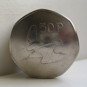 May include: A silver-colored heptagonal coin featuring a relief design of a bird in flight on the face. The text 50P is clearly visible in the upper right quadrant of the coin, indicating its denomination as a fifty pence piece.