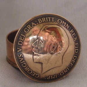 May include: A round copper-toned metal box with a lid featuring a relief portrait of King George V in profile. The text GEORGIVS V DEI GRA: BRITT: OMN: REX FID: DEF: IND: IMP: is embossed around the edge of the lid, which rests on a cylindrical base.