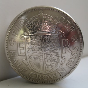 May include: A silver Half Crown coin from 1955. The coin features a detailed design of a crown, shield, and text around the edge. The words "HALF CROWN" and "1955" are visible. The coin has a detailed edge.