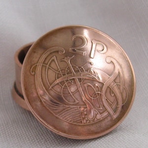 May include: A copper-colored ring with a coin-like top. The coin features the number "2P" and an intricate Celtic design. The ring's band is visible, suggesting it's a wearable piece of jewelry.