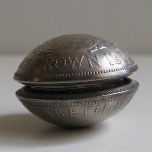 May include: A silver coin container, split open. The top half reads "HALF CROWN 1963" and the bottom half reads "ELIZABETH II". The container is round with a textured edge. A vintage coin holder.