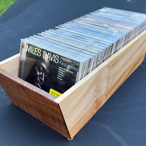 May include: A rectangular wooden storage bin with angled sides, crafted from light-toned wood with a visible grain pattern. The bin is filled with a row of clear plastic CD cases, with the Miles Davis Kind of Blue album visible at the front.