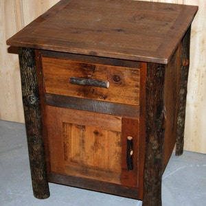 May include: Rustic wooden nightstand with a drawer and cabinet door. The nightstand is constructed from various shades of brown wood, with log accents on the legs and sides. The drawer and door have twig-like handles. The top is a darker shade of brown.