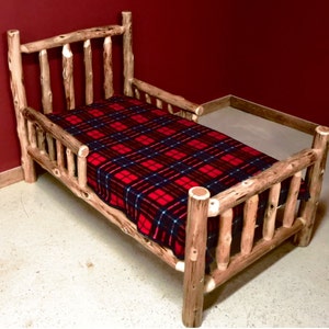 May include: A wooden toddler bed with a red and black plaid blanket. The bed has a high headboard and footboard, and a safety rail on one side.