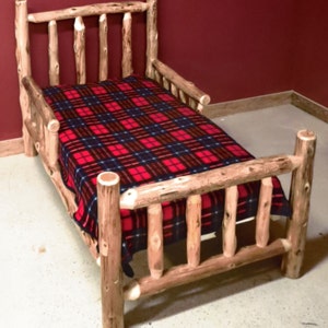 May include: A wooden toddler bed with a red and black plaid blanket. The bed has a headboard and footboard with a slatted design. The bed is made of natural wood with a rustic finish.