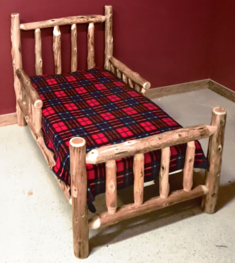May include: A wooden toddler bed with a red and black plaid blanket. The bed has a headboard and footboard with a slatted design. The bed is made of natural wood with a rustic finish.