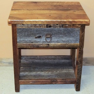 May include: A rustic wooden end table with a single drawer and a lower shelf. The table is made of reclaimed wood with a natural finish. The drawer has a metal ring pull.