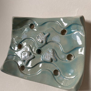 May include: A light blue ceramic soap dish with a wavy design and three fish shapes. The dish has drainage holes.