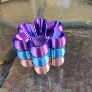 Puede incluir: Un objeto con forma de flor de tres niveles con un acabado suave y brillante. La capa superior es de un color morado vibrante, la capa intermedia es de un azul cian brillante y la capa inferior es de un color cobre metálico suave.