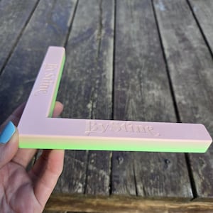 Op de afbeelding: Een groen, roze en wit plastic hoekstuk met de tekst "By Slime" erop gegraveerd.