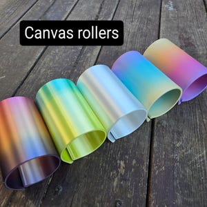 May include: Five canvas rollers in a range of colours, including purple, orange, green, blue, and silver. The rollers are displayed on a weathered wooden surface. The text "Canvas rollers" is in a black rectangle.