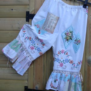 Könnte beinhalten: Weiße Caprihose mit floralen Stickereien und Patchwork-Details. Die Hose hat einen gerüschten Saum mit Spitzenbesatz und ist mit Flicken aus floralem Stoff und einem Vintage-Papierelement verziert. Die Hose hängt an einem schwarzen Kleiderbügel.