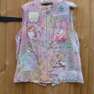 Peut inclure: Un gilet sans manches dans des tons rose et crème avec un motif floral, des plis verticaux et une bordure en dentelle. Le vêtement est orné de divers empiècements en tissu, incluant des images de lapins dans des tasses et des compositions florales.