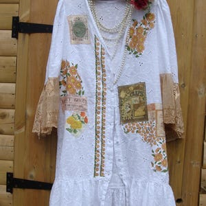 May include: White eyelet duster coat with floral and vintage label appliques. It has lace sleeves, a ruffled hem, and a beaded necklace. The button-down coat features a decorative floral accent on the shoulder.