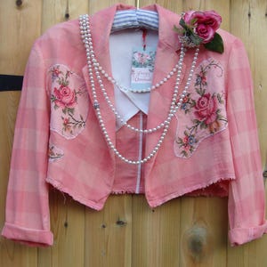 Puede incluir: Chaqueta rosa a cuadros con bordados florales y collar de perlas. La chaqueta tiene mangas enrolladas y un cuello blanco. Una rosa rosa y una etiqueta "Pretty Unusual" están adjuntas.