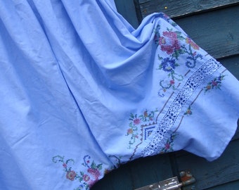 Op de afbeelding: Een lichtblauwe rok met geborduurde bloemenmotieven en een kantversiering. De rok hangt aan een hanger tegen een blauwe houten achtergrond.