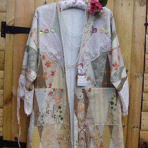 Può includere: Cappotto patchwork con motivi floreali e geometrici nei toni del beige, crema e marrone. Il cappotto presenta dettagli in pizzo, bottoni decorativi e una collana di perle. È attaccata una rosa rosa e un'etichetta "Pretty Unusual".