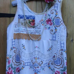 Puede incluir: Vestido blanco sin mangas con bordado floral de punto de cruz en rosa, azul y verde. El vestido presenta ribete de encaje, parches con imágenes impresas y un collar de perlas. Una rosa rosa adorna la percha.