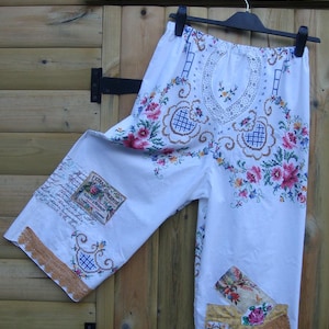 Könnte beinhalten: Weiße, bestickte Caprihose mit floralen und geometrischen Mustern. Auf die Hose sind Aufnäher mit Text und floralen Designs genäht. Die Hose hängt an einem schwarzen Kleiderbügel vor einem hölzernen Hintergrund.