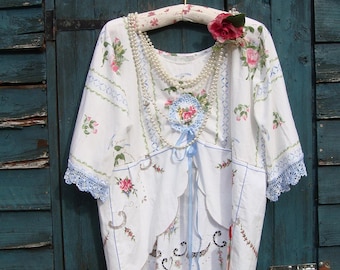 Puede incluir: Top blanco bordado con flores rosas y hojas verdes, con ribete de encaje azul y dobladillo festoneado. Tiene una cinta azul y un collar de perlas. Una rosa roja adorna la percha.