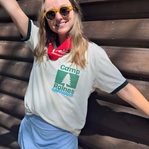 May include: A person wearing a white t-shirt with a green and blue logo that says 'Camp Raines' and a red bandana around their neck. They are wearing yellow sunglasses and a blue skirt.